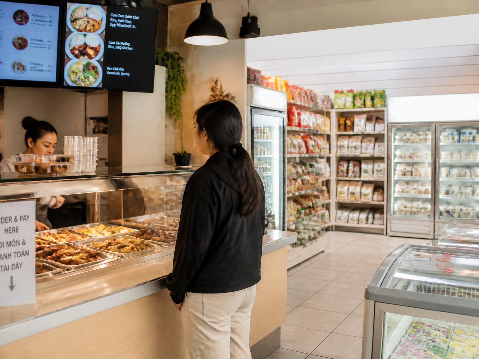 Customer waiting at a hot food counter with retail shelves, chiller and freezer behind